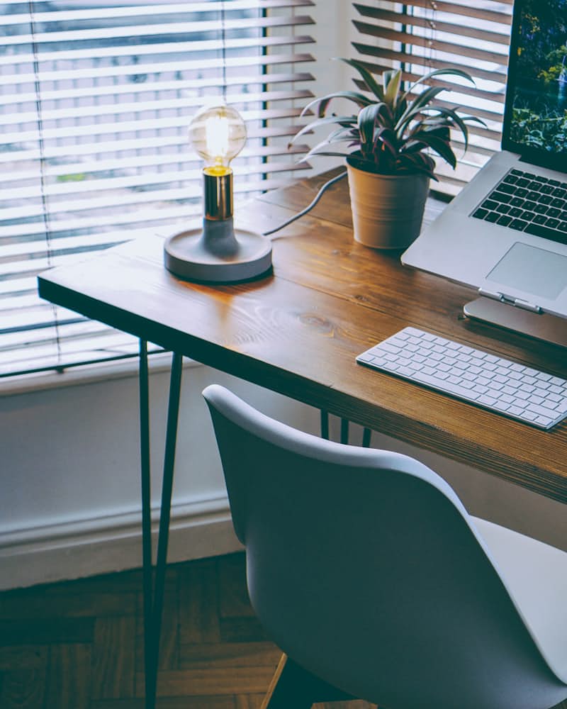 A wooden desk by a shuttered window with a laptop, a keyboard, a bulb lamp and a potted plant
