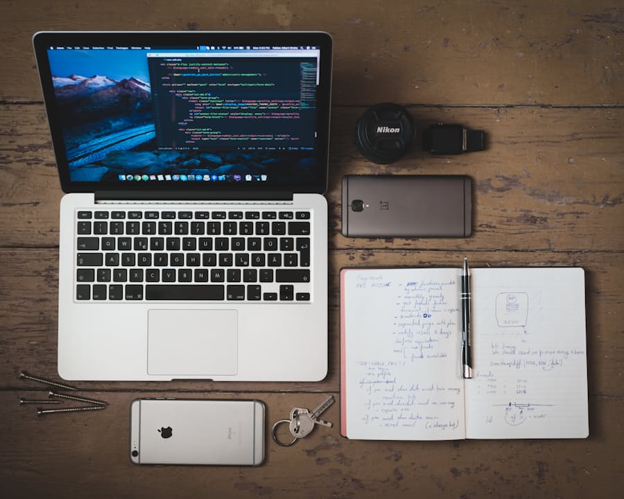 A wooden desk from above with an open laptop, a handwritten notebook, a phone and a camera lens
