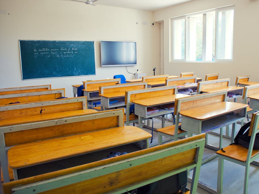 An empty classroom with rows of wooden desks, a blackboard and a wall-mounted screen
